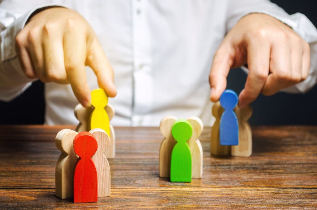SEO Hannover - Eine Person ordnet Holzfiguren auf einem Tisch an. Die Figuren haben die Form von Menschen und sind in verschiedenen Farben bemalt: rot, grün, gelb und blau. Die Person verwendet beide Hände, um die Figuren zu positionieren, was auf eine ordnende Tätigkeit hindeutet. Local SEO Agentur Hannover.