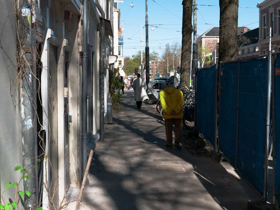 Suchmaschinenoptimierung Hannover - Eine Person in einer gelben Jacke geht eine schmale Straße entlang, die links von Gebäuden und rechts von einem blauen Bauzaun gesäumt ist. Auf dem Gehweg liegen Schattenflecken von den Bäumen entlang der Straße. Weiter unten auf dem Gehweg sind weitere Personen zu sehen. Local SEO Agentur Hannover.