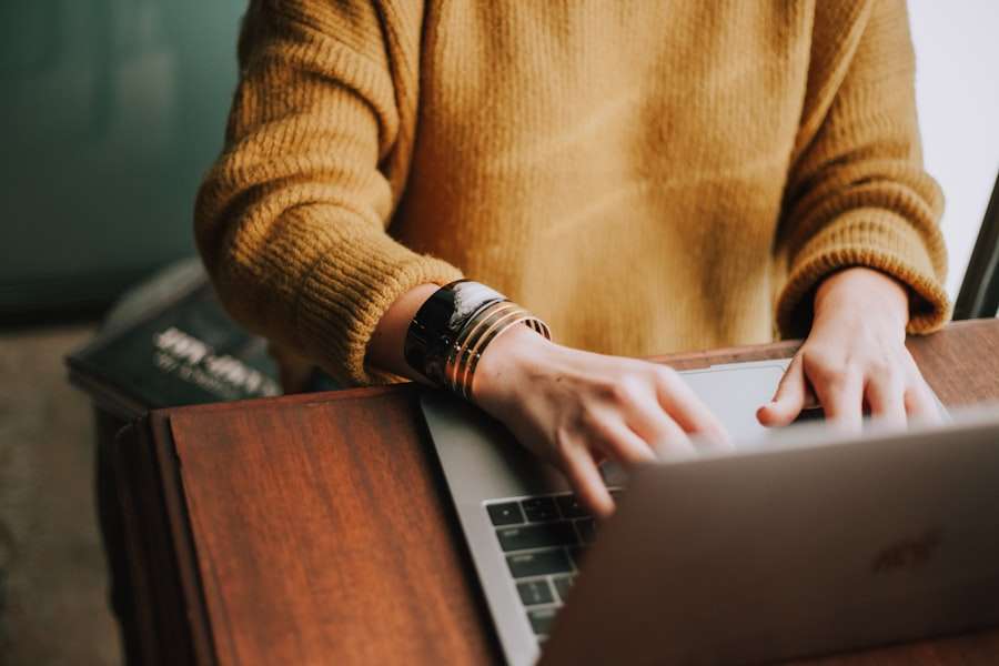 SEO Hannover - Eine Person in einem senfgelben Pullover und mehreren Armbändern tippt auf einem Laptop, der auf einem Holzschreibtisch steht. Der Hintergrund ist leicht verschwommen, wodurch der Fokus des Motivs auf dem Bildschirm hervorgehoben wird. Local SEO Agentur Hannover.