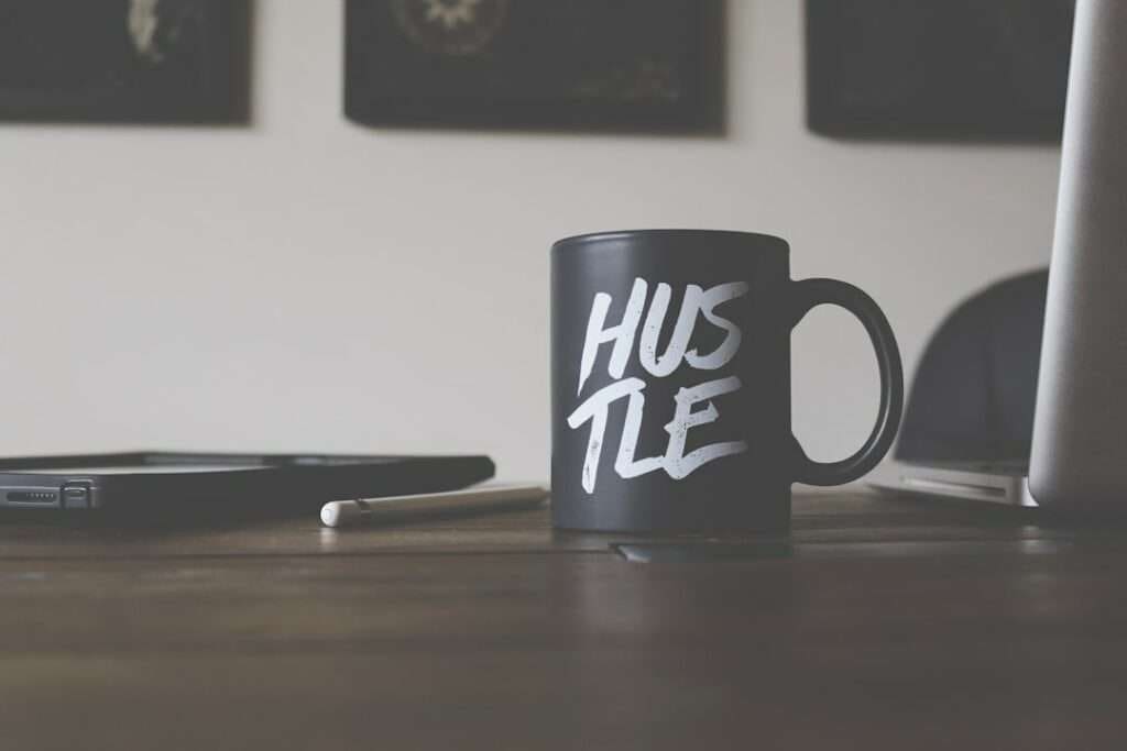 Suchmaschinenoptimierung Hannover - Eine schwarze Kaffeetasse mit dem Wort „HUSTLE“ in weißen Buchstaben steht auf einem Holztisch. Neben der Tasse liegen ein Stift und ein geschlossener Laptop, ideal, wenn Sie eine Webvisitenkarte erstellen müssen. Ein weiteres elektronisches Gerät ist teilweise im schwach beleuchteten Hintergrund sichtbar. Local SEO Agentur Hannover.