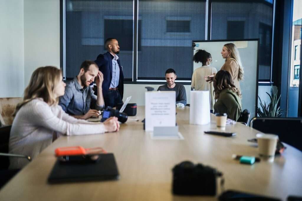 Suchmaschinenoptimierung Hannover - Eine Gruppe von sieben Personen hat sich in einem modernen Büro-Besprechungsraum einer Suchmaschinenoptimierungsagentur versammelt. Einige sitzen mit Laptops und Notizbüchern an einem langen Holztisch, andere stehen und unterhalten sich an einem Whiteboard. Im Hintergrund sind große Fenster mit Jalousien zu sehen. Local SEO Agentur Hannover.
