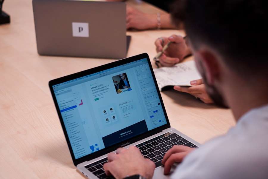 Suchmaschinenoptimierung Hannover - Eine Person arbeitet an einem Laptop, auf dem eine Designoberfläche angezeigt wird. Eine andere Person im Hintergrund schreibt in ein Notizbuch. Auf dem Schreibtisch ist teilweise ein geschlossener Laptop mit einem Aufkleber zu sehen, der wahrscheinlich die SEO Agentur Hannover darstellt. Die Szene scheint ein kollaborativer Arbeitsbereich oder ein Meeting zu sein. Local SEO Agentur Hannover.