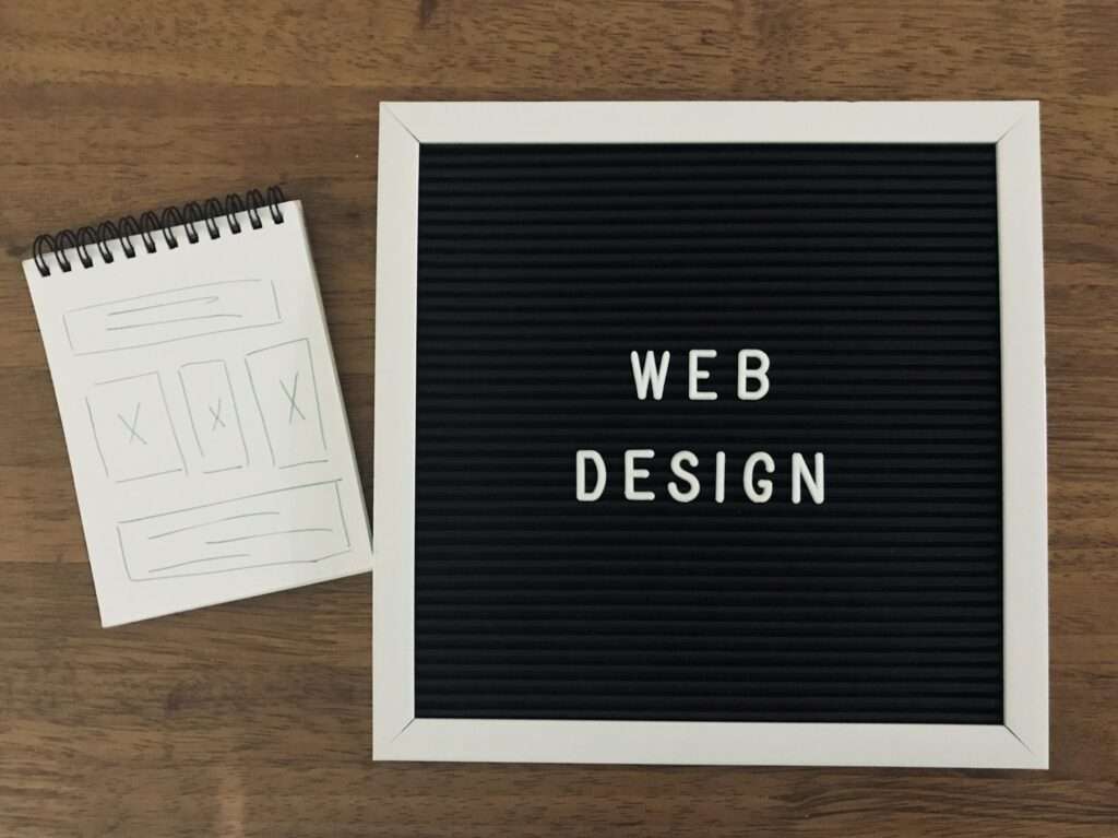 SEO Hannover - Rechts ist eine schwarze Buchstabentafel mit weißen Buchstaben, auf denen „WEB DESIGN“ steht, angebracht. Links liegt ein kleines Spiralnotizbuch aufgeschlagen, auf dem skizzierte Website-Layout-Ideen mit einem detaillierten Kopfmenü zu sehen sind. Beide Elemente liegen auf einer Holzoberfläche. Local SEO Agentur Hannover.