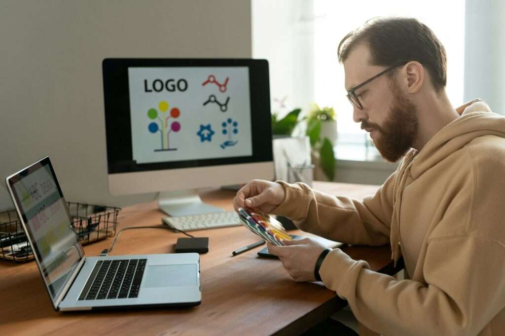 SEO Hannover - Eine Person mit Brille, Bart und Kapuzenpullover sitzt an einem Schreibtisch und bedient ein Tablet. Auf zwei Computerbildschirmen wird ein Logo-Design und ein Text mit der Aufschrift „Logo erstellen lassen“ angezeigt. Auf dem Schreibtisch stehen Büromaterial und eine Pflanze, während Sonnenlicht durch das Fenster fällt. Local SEO Agentur Hannover.