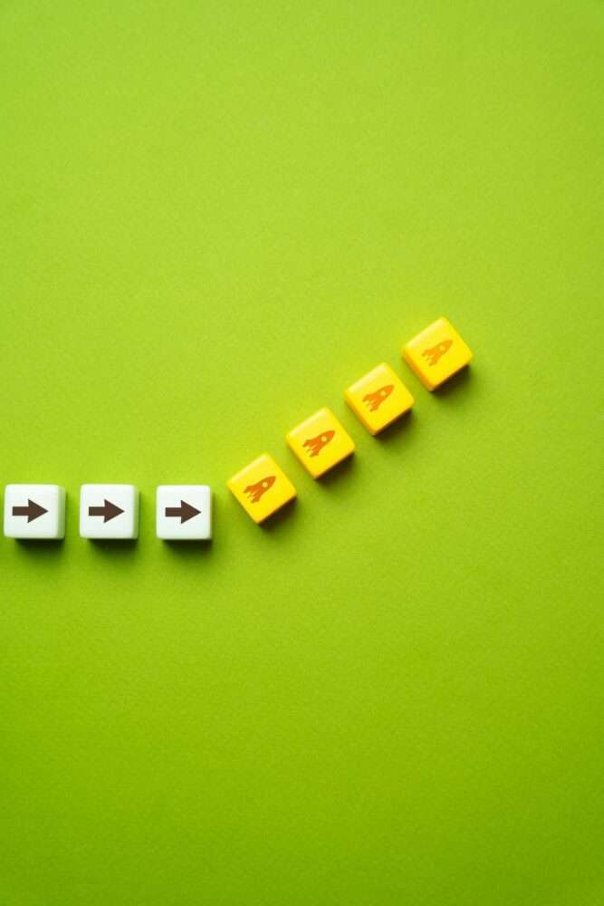 Webdesign Hannover - Eine diagonale Linie aus weißen und gelben Würfeln auf grünem Hintergrund veranschaulicht geschickt die strategischen Schritte bei der Google My Business Optimierung. Die weißen Würfel sind mit schwarzen Pfeilsymbolen versehen, die nach rechts zeigen, während die gelben Würfel mit Raketensymbolen geschmückt sind. Local SEO Agentur Hannover.
