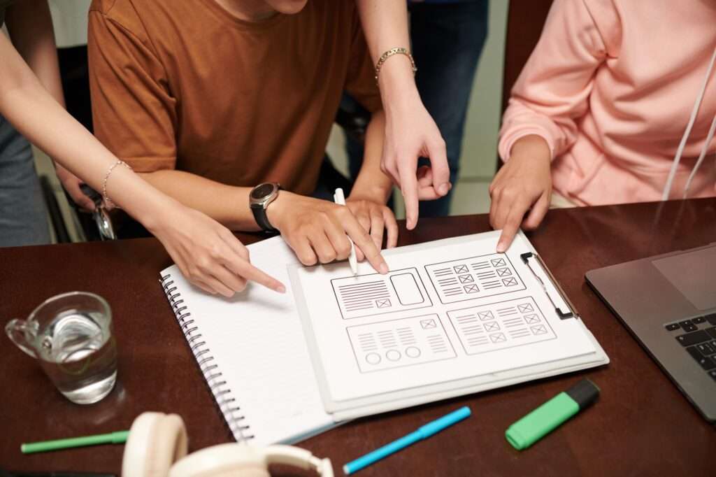 Webdesign Hannover - Eine Gruppe von Leuten sitzt um einen Tisch herum und zeigt auf ein digitales Drahtmodell auf einem Klemmbrett. Sie scheinen zu diskutieren, was auf einer Website enthalten sein muss. Auf dem Tisch liegen die wichtigsten Dinge herum: ein Notizbuch, ein Glas Wasser, Stifte und ein Laptop. Local SEO Agentur Hannover.