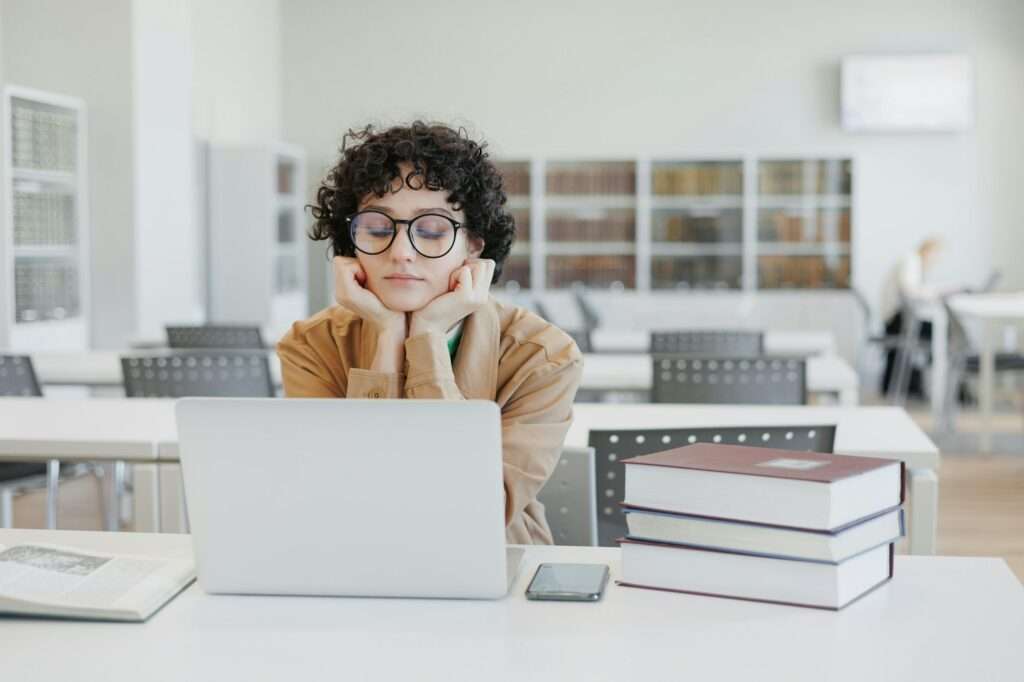 Webdesign Hannover - Eine Person mit lockigem Haar und Brille sitzt an einem Bibliothekstisch und stützt ihr Kinn auf die Hände vor einem geöffneten Laptop, auf dem eine barrierefreie Webseite angezeigt wird. Um sie herum sind Stapel von Büchern, ein Smartphone und ein weiteres Buch. Im Hintergrund stehen Regale voller Bücher. Local SEO Agentur Hannover.