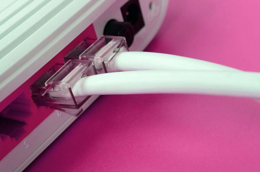 Webdesign Hannover - Nahaufnahme von zwei weißen Ethernet-Kabeln, die vor einem rosa Hintergrund an der Rückseite eines grauen, barrierefreien Routers eingesteckt sind. Local SEO Agentur Hannover.