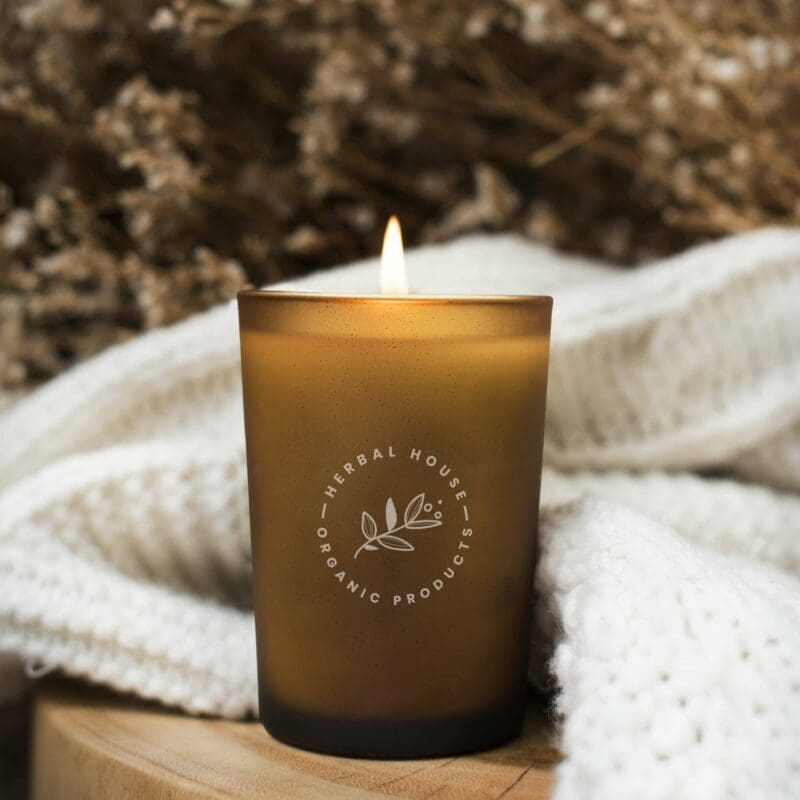 Webdesign Hannover - Eine brennende braune Kerze in einem Milchglashalter mit dem Logo „Herbal House Organic Products“ steht elegant auf einer Holzoberfläche. Mit leichtem Strickstoff drapiert und mit getrockneten Blumen akzentuiert, ist sie die perfekte Kulisse, um Ihr eigenes, einzigartiges Design zu präsentieren. Logo erstellen lassen für persönliches Flair. Local SEO Agentur Hannover.