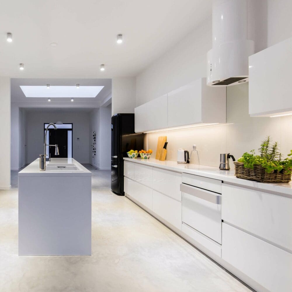 SEO Hannover - Diese moderne Küche, perfekt für Immobilienfotografie, besticht durch weiße Schränke und eine Kücheninsel. Topfpflanzen und kleine Haushaltsgeräte schmücken die Oberflächen, während in der Ecke ein schwarzer Kühlschrank steht. Deckenleuchten und ein Oberlicht erhellen den Raum hervorragend. Local SEO Agentur Hannover.