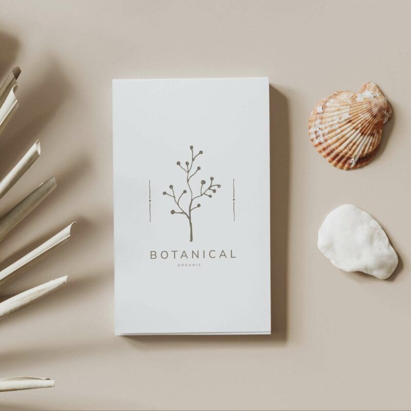 Webdesign Hannover - Eine weiße Broschüre mit botanischem Design und dem Text „BOTANICAL ORGANIC“ unterstreicht auf elegante Weise die Kunstfertigkeit des Logodesigns. Sie ruht auf einer beigen Oberfläche, begleitet von weißen Stöcken, einer Muschel und einem weißen Stein. Local SEO Agentur Hannover.