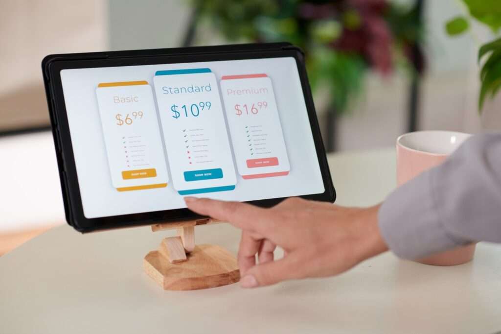 Webdesign Hannover - Eine Person zeigt auf ein Tablet, auf dem Abonnements für die Website-Wartung angezeigt werden. Auf dem Bildschirm sind drei Optionen zu sehen: Basic für 6,99 $, Standard für 10,99 $ und Premium für 16,99 $, jede mit einer eigenen Liste der Funktionen darunter. Im Hintergrund steht eine rosa Tasse, die die gemütliche Atmosphäre der Szene verstärkt. Local SEO Agentur Hannover.