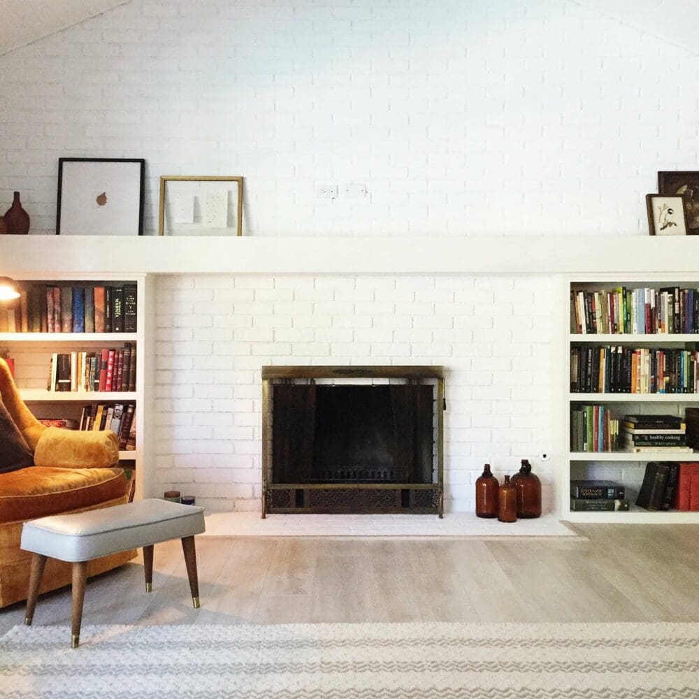SEO Hannover - Ein gemütliches Wohnzimmer präsentiert einen weißen gemauerten Kamin, ideal für Immobilienfotografie, mittig an der Wand und flankiert von eingebauten Bücherregalen voller Bücher und Dekoration. Ein orangefarbener Sessel und eine graue Bank stehen auf einem hellen Holzboden mit einem gemusterten Teppich. Local SEO Agentur Hannover.