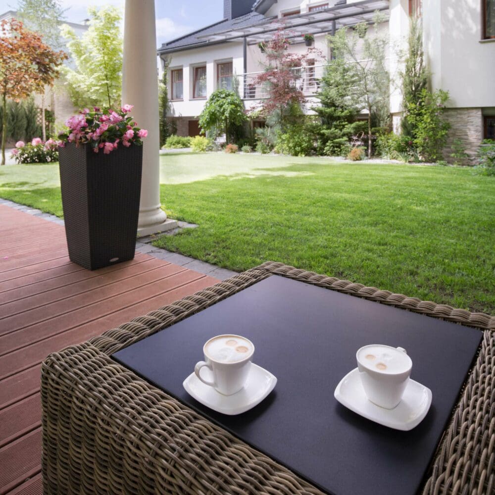 SEO Hannover - Zwei Cappuccinotassen stehen auf einem quadratischen Korbtisch auf einer Terrasse, perfekt für ein Immobilienfotografie-Shooting. Der Platz blickt auf einen gepflegten Garten mit üppigem Gras, Bäumen und leuchtenden Blumen. In der Nähe steht eine große Pflanze in einem Topf, im Hintergrund ist ein Gebäude mit großen Fenstern zu sehen. Local SEO Agentur Hannover.