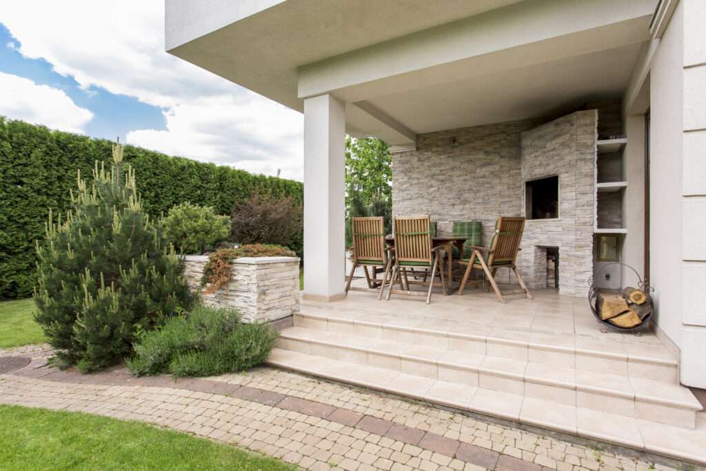 SEO Hannover - Eine moderne Außenterrasse, perfekt für das Portfolio eines Immobilienfotografen, verfügt über ein Esszimmer mit sechs Holzstühlen und einem Tisch auf einem gefliesten Boden. Es gibt einen eingebauten Grillbereich aus Stein, einen Stapel Brennholz und umgebendes Grün. Ein gepflasterter Weg führt zu diesem einladenden Platz. Local SEO Agentur Hannover.