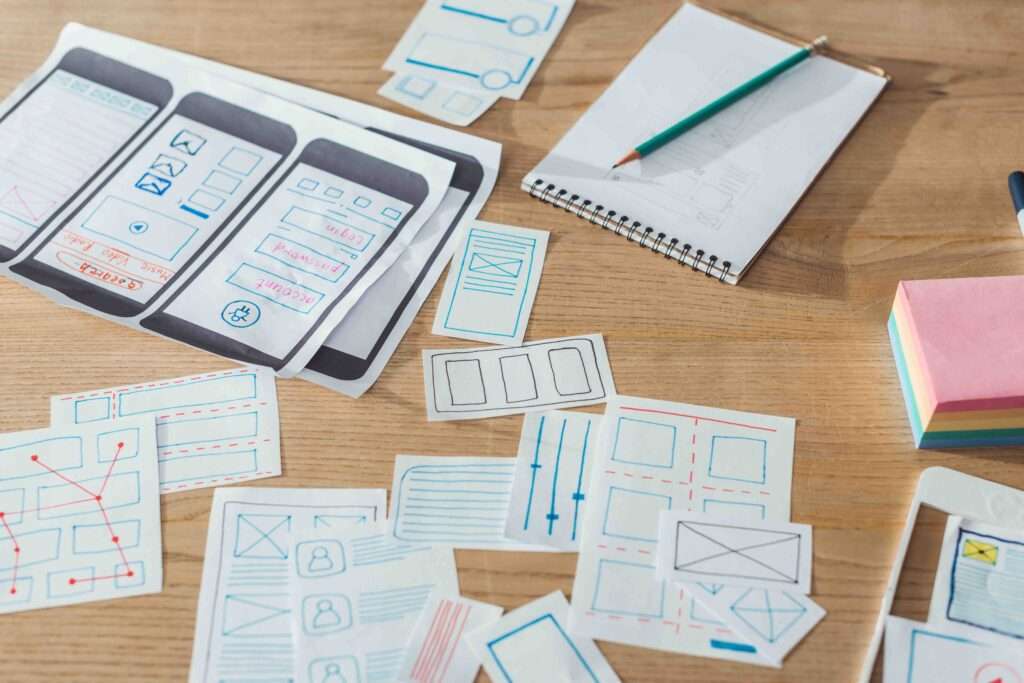 Webdesign Hannover - Ein Holztisch voller Papierskizzen von Designs für mobile Benutzeroberflächen ähnelt der Struktur einer HTML-Sitemap. Ein Bleistift liegt auf einem weißen Notizbuch neben einem Stapel bunter Haftnotizen, während die Skizzen Drahtgitterlayouts, Symbole und Pfeile zeigen. Local SEO Agentur Hannover.