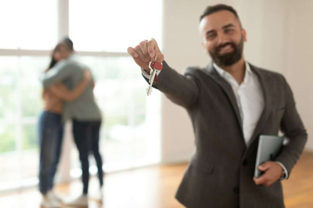 SEO Hannover - Ein Mann im Anzug lächelt und hält Schlüssel in die Kamera, was an einen erfolgreichen Deal auf einer Webseite für Immobilienmakler erinnert. Im Hintergrund umarmt sich ein Paar in einem verschwommenen Raum mit großen Fenstern. Local SEO Agentur Hannover.