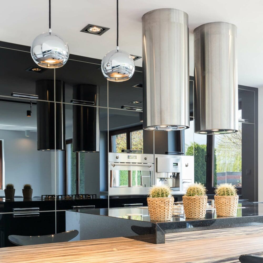 SEO Hannover - Moderne Küche mit einem eleganten Design aus Schwarz und Edelstahl, perfekt eingefangen durch Immobilienfotografie. Zwei große Dunstabzugshauben aus Metall hängen über einer Arbeitsplatte mit drei kleinen Kakteen in Töpfen, während die reflektierenden Oberflächen und Einbaugeräte des Raums den zeitgenössischen Look vervollständigen. Local SEO Agentur Hannover.