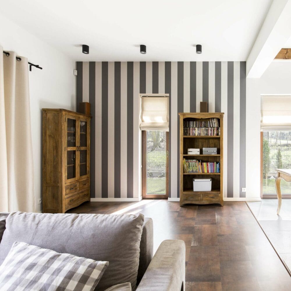 SEO Hannover - Ein Wohnzimmer, das im Stil der Immobilienfotografie präzise festgehalten wurde, zeigt ein graues Sofa mit einem karierten Kissen. Die gestreifte Akzentwand wird von zwei hohen, mit Büchern gefüllten Schränken eingerahmt. Beigefarbene Jalousien zieren die beiden Fenster und Licht fällt von draußen auf den dunklen Holzboden. Local SEO Agentur Hannover.
