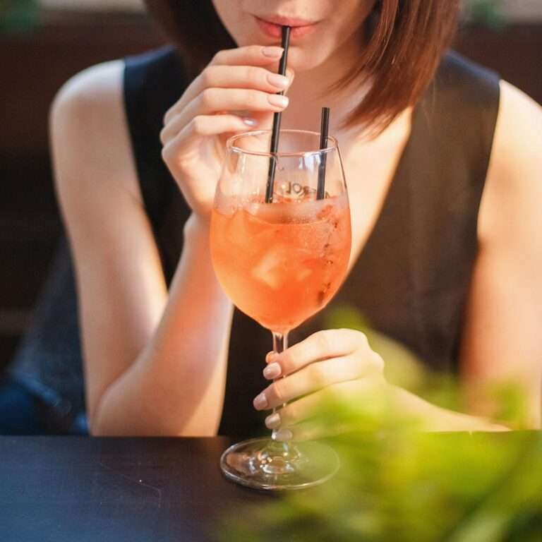 SEO Hannover - Eine Person in einem ärmellosen schwarzen Oberteil sitzt an einem Tisch und nippt mit einem schwarzen Strohhalm an einem orangefarbenen Getränk mit Eis aus einem Stielglas. Die Szene, die sich perfekt zur Verbesserung von SEO für Bilder eignet, zeigt im Vordergrund leicht verschwommenes Grün, was der lebendigen Umgebung Tiefe verleiht. Local SEO Agentur Hannover.