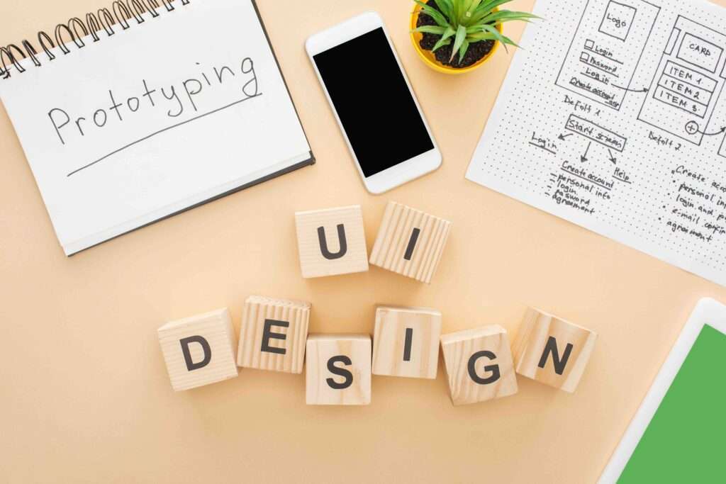 Webdesign Hannover - Ein Arbeitsplatz mit einem Notizbuch mit der Aufschrift „Prototyping“, einem Smartphone, einer kleinen Pflanze und einem Blatt mit UI-Design-Skizzen. In der Mitte sind Holzklötze mit der Aufschrift „UI DESIGN“ angeordnet, die die wesentlichen Elemente widerspiegeln, die auf einer Website nicht fehlen dürfen. Local SEO Agentur Hannover.