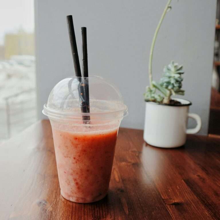 SEO Hannover - Ein Plastikbecher mit einem rosa Smoothie steht auf einem Holztisch und verkörpert den perfekten Moment für SEO für Bilder. Der durchsichtige Kuppeldeckel hält zwei schwarze Strohhalme, während im Hintergrund ein weißer Becher mit einer Sukkulente die ruhige Innenatmosphäre neben einem großen Fenster unterstreicht. Local SEO Agentur Hannover.