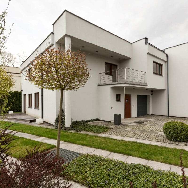 SEO Hannover - Ein modernes, zweistöckiges weißes Haus mit Flachdach und Balkon, perfekt für Immobilienfotografie. Der Eingang verfügt über eine Holztür, rechts befindet sich ein Garagentor. Ein kleiner Baum, Sträucher und ein gepflasterter Gehweg schmücken den Vorgarten, während darüber ein bedeckter Himmel thront. Local SEO Agentur Hannover.