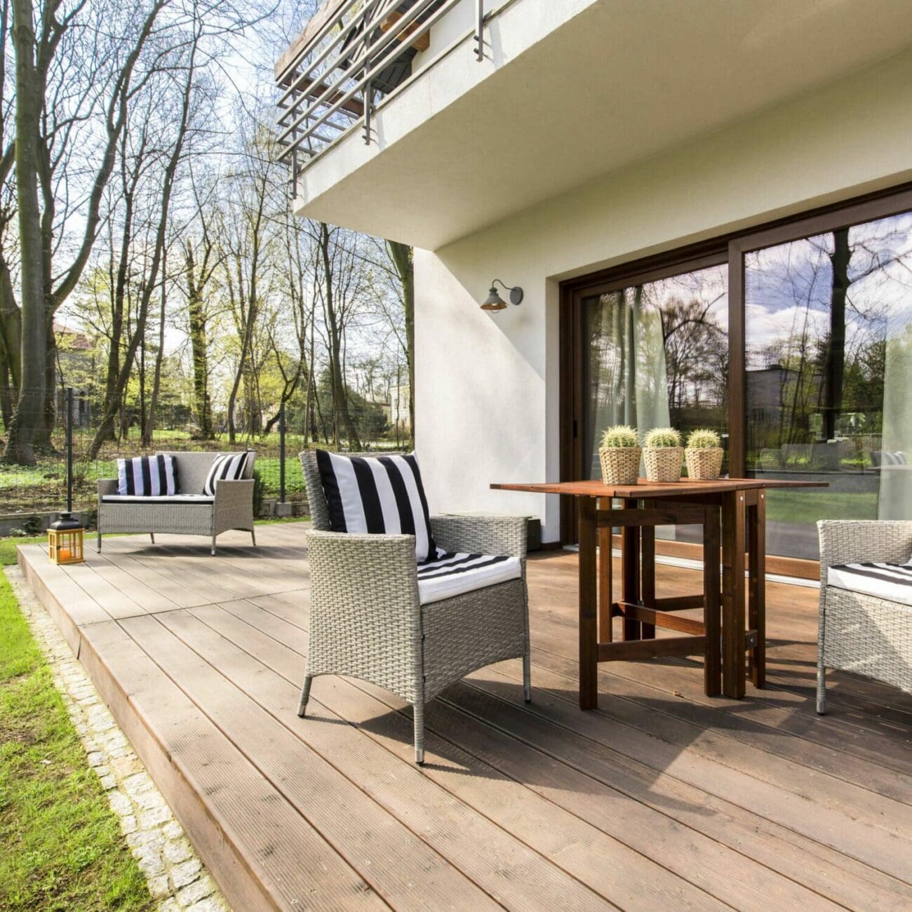 SEO Hannover - Auf einer Holzterrasse stehen zwei graue Korbstühle mit gestreiften Kissen gegenüber einem kleinen Tisch, der mit Kakteen in Töpfen geschmückt ist, perfekt für die Aufnahme in Immobilienfotografie. Eine Glasschiebetür führt hinein, während Bäume und Gras die Terrasse friedlich umgeben. Local SEO Agentur Hannover.