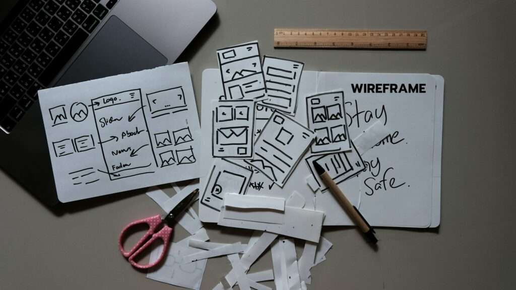 Webdesign Hannover - Auf einem Schreibtisch stehen ein Laptop, ein Lineal, eine Schere und Drahtgitter-Skizzen. Papierausschnitte von Web-Layouts liegen verstreut neben Zettelchen mit der Aufschrift „Bleiben Sie zu Hause. Bleiben Sie gesund.“ Inmitten des Chaos heben verschiedene Arten von Sitemaps unterschiedliche Site-Strukturen hervor. Local SEO Agentur Hannover.