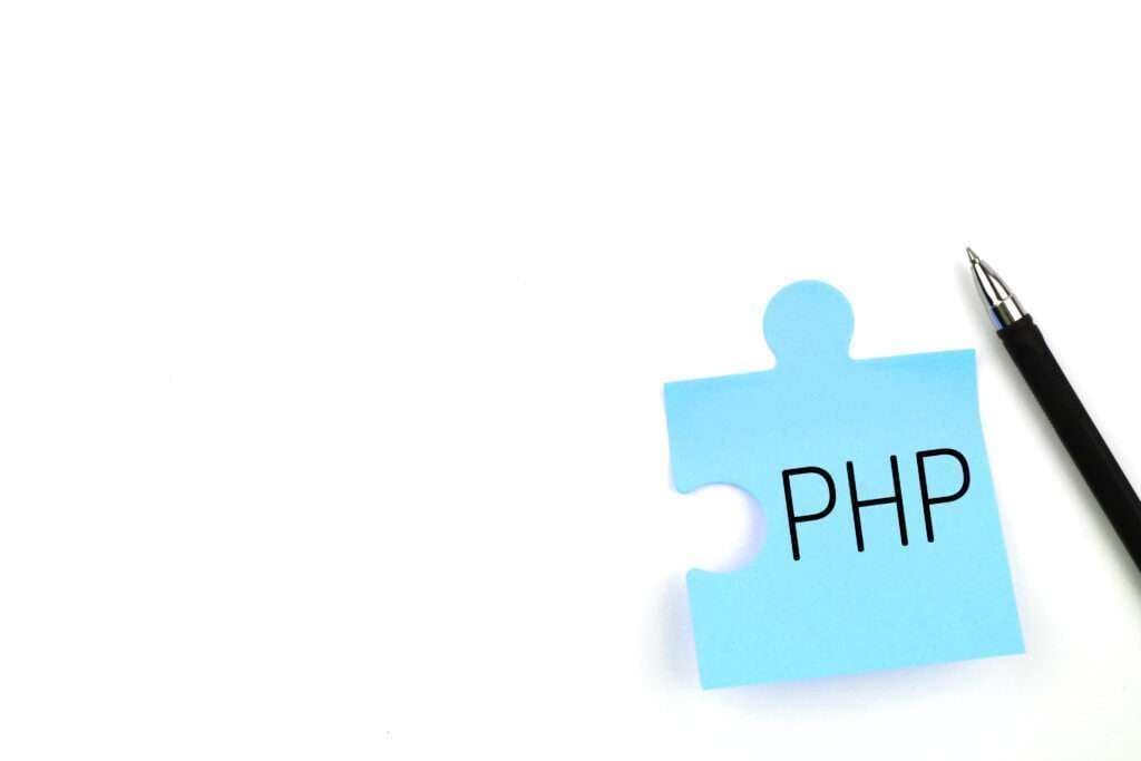 Webdesign Hannover - Ein blaues Puzzleteil mit den Buchstaben „PHP“ befindet sich neben einem schwarzen Stift auf weißem Hintergrund und fängt die Essenz von „Was ist PHP einfach erklärt?“ in einer einfachen visuellen Metapher ein. Local SEO Agentur Hannover.