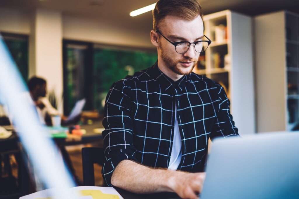 Webdesign Hannover - Ein Mann mit Brille sitzt an einem Schreibtisch und konzentriert sich darauf, auf seinem Laptop „Wie finde ich meinen PageRank bei Google?“ herauszufinden. Er trägt ein schwarzes Hemd mit einem weißen Gittermuster. Im Hintergrund arbeitet eine andere Person an einem Tisch in einem gut beleuchteten Raum mit Bücherregalen. Local SEO Agentur Hannover.