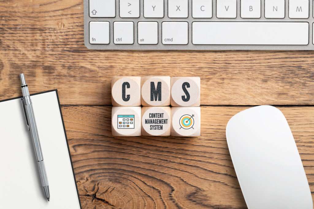 Webdesign Hannover - Holzklötze bilden das Wort „CMS“ auf einem Holzschreibtisch, umgeben von Symbolen, die einen Zeitplan und ein Ziel darstellen und die WordPress-Wartung veranschaulichen. In der Nähe finden Sie eine Tastatur, eine Maus, einen Stift und einen Notizblock, die zum Einsatz bereit sind. Local SEO Agentur Hannover.
