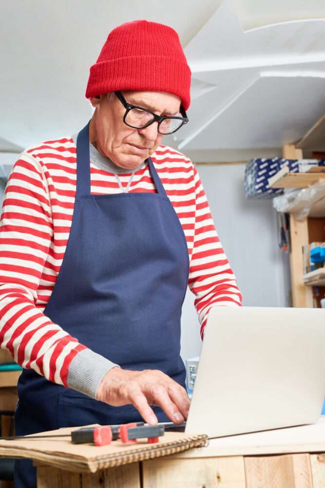 Webdesign Hannover - Ein älterer Erwachsener, der einem Handwerker in einer roten Mütze und einem gestreiften Pullover ähnelt, arbeitet in seiner Werkstatt an einem Laptop. Er trägt eine Brille und eine marineblaue Schürze. In der Nähe liegen ein Maßband und ein Bleistift auf dem Holztisch und deuten auf Pläne für die Erstellung ansprechender Webseiten hin. Im Hintergrund sind Regale zu sehen. Local SEO Agentur Hannover.