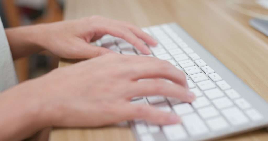 Webdesign Hannover - Hände tippen auf einer weißen Computertastatur auf einem Holzschreibtisch. Das Bild konzentriert sich auf die Finger, wobei Tastatur und Schreibtischoberfläche deutlich zu sehen sind. Es regt zum Nachdenken an: Sind Schlüsselwörter in diesem digitalen Zeitalter, in dem Bilder mehr sagen als Worte, noch wichtig? Local SEO Agentur Hannover.