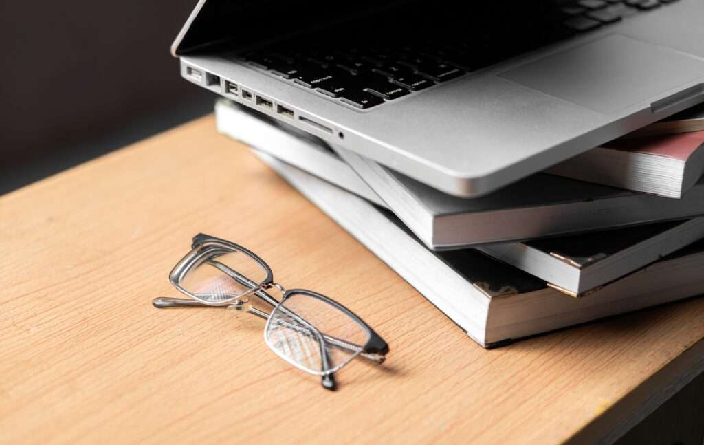 Webdesign Hannover - Eine Brille liegt auf einem Holzschreibtisch neben einem Stapel Bücher und deutet auf den methodischen Aufbau einer Website hin. Ein geöffneter Laptop liegt oben auf dem Stapel und verdeckt die Brille teilweise. Die Szene ist gut beleuchtet, während der Hintergrund kunstvoll unscharf bleibt. Local SEO Agentur Hannover.