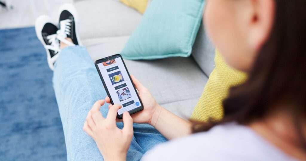 Webdesign Hannover - Eine Person sitzt mit einem Smartphone auf einer Couch. Sie trägt blaue Jeans und schwarze Turnschuhe mit weißen Schnürsenkeln. Auf dem Telefonbildschirm wird eine App mit einem Dropdown-Menü mit mehreren Bildern und Optionen angezeigt. Im Hintergrund sorgen ein blauer Teppich und bunte Kissen für eine gemütliche Atmosphäre. Local SEO Agentur Hannover.