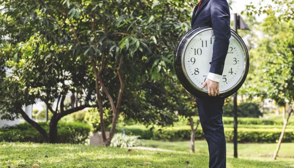 Webdesign Hannover - Eine Person im Anzug steht auf dem Rasen in einem Park und hält eine große runde Wanduhr unter dem Arm. Sie denkt vielleicht über die Verweildauer der Zeit nach. Bäume und Grün rahmen die Szene ein, während die Zifferblätter 9:10 anzeigen. Local SEO Agentur Hannover.