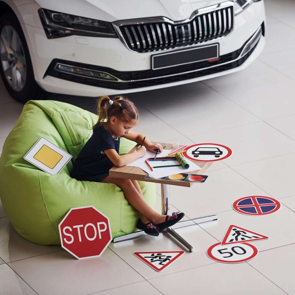 Webdesign Hannover - Ein junges Mädchen sitzt auf einem grünen Sitzsack und malt an einem kleinen Tisch, umgeben von verschiedenen Verkehrsschildern. Hinter ihr parkt ein weißes Auto im Innenbereich, was auf eine lehrreiche Veranstaltung hindeutet – perfekt für eine Präsentation von Autohaus-Webseiten erstellen lassen. Local SEO Agentur Hannover.