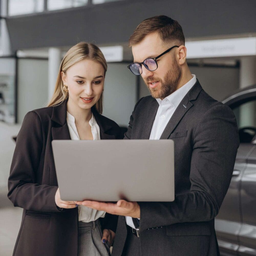 Webdesign Hannover - Ein Mann und eine Frau in Geschäftskleidung stehen zusammen in einem Autohaus und schauen auf einen Laptop, den der Mann in der Hand hält. Vielleicht diskutieren sie, wie sie Autohaus-Webseiten erstellen können, während im Hintergrund mehrere Autos ausgestellt sind. Local SEO Agentur Hannover.