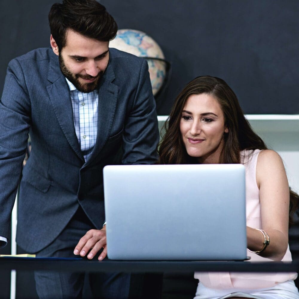 Webdesign Hannover - Ein Mann im Anzug steht neben einer sitzenden Frau. Beide schauen auf einen Laptop-Bildschirm und diskutieren möglicherweise darüber, wie man Autohaus-Webseiten erstellen kann. Im Hintergrund sind eine Tafel und ein Globus zu sehen. Local SEO Agentur Hannover.