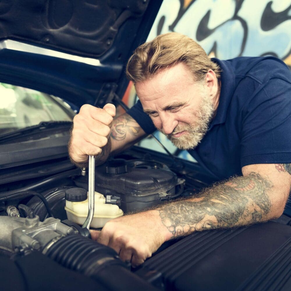 Webdesign Hannover - Ein Mann mit Tattoos und Bart arbeitet konzentriert mit einem Schraubenschlüssel an einem Automotor. Er trägt ein marineblaues Hemd, hinter ihm eine Graffiti-Wand – perfekt für die Präsentation eines Autohaus-Webseiten-erstellen-lassen-Projekts. Local SEO Agentur Hannover.