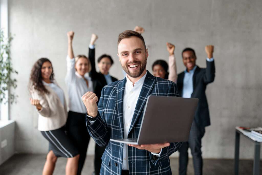 Webdesign Hannover - Vorne steht ein Mann im karierten Jackett, hält einen Laptop in der Hand und lächelt, während hinter ihm fünf Kollegen feiern – und so in einer Büroumgebung ihre Erfolge mit Google Maps SEO Hannover präsentieren. Local SEO Agentur Hannover.