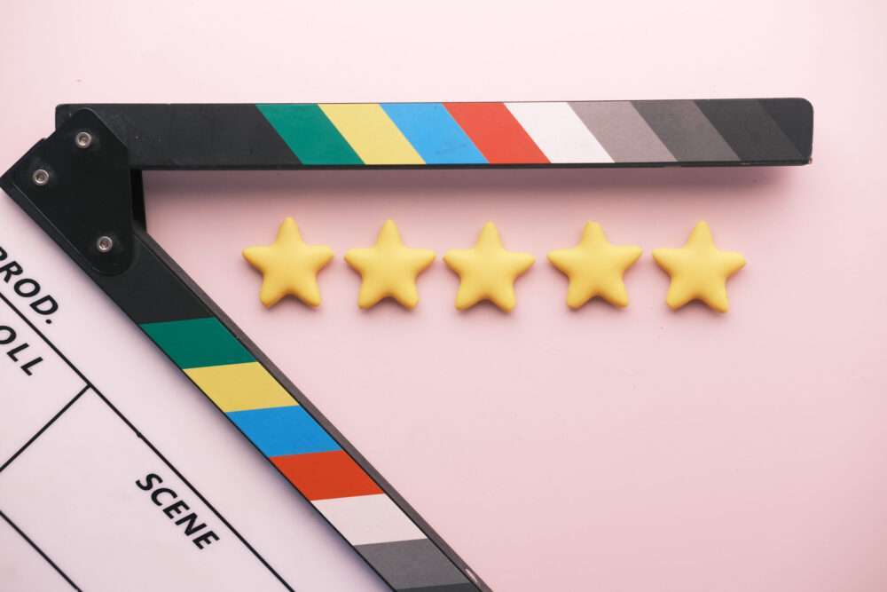 Webdesign Hannover - Eine Filmklappe mit farbigen Streifen ist auf einem rosa Hintergrund geöffnet. Davor deuten fünf gelbe Sterne auf eine Fünf-Sterne-Bewertung hin, ähnlich wie Top-Google-Rezensionen von zufriedenen Zuschauern. Local SEO Agentur Hannover.