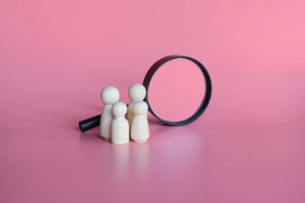 Webdesign Hannover - Vier Holzfiguren stehen vor einem aufrechten Vergrößerungsglas auf rosafarbenem Hintergrund und erinnern an Menschen, die genau unter die Lupe genommen werden - so wie ein SEO Website Check jedes Detail für die Online-Optimierung prüft. Local SEO Agentur Hannover.