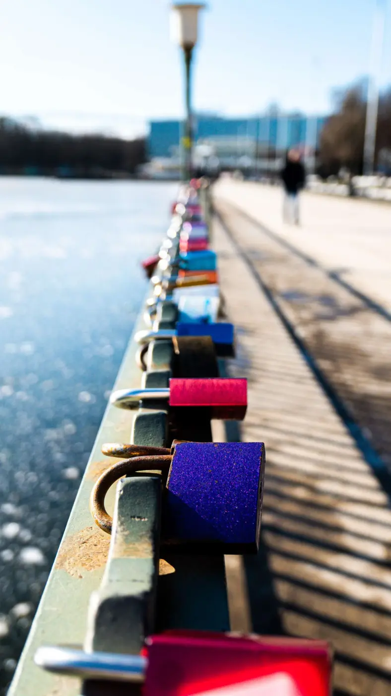 Webdesign Hannover - Eine Reihe von bunten Vorhängeschlössern, die an einem grünen Metallgeländer neben einem Fluss befestigt sind, symbolisiert wordpress sicherheit, mit einem Gehweg und verschwommenen Menschen im Hintergrund an einem sonnigen Tag. Local SEO Agentur Hannover.