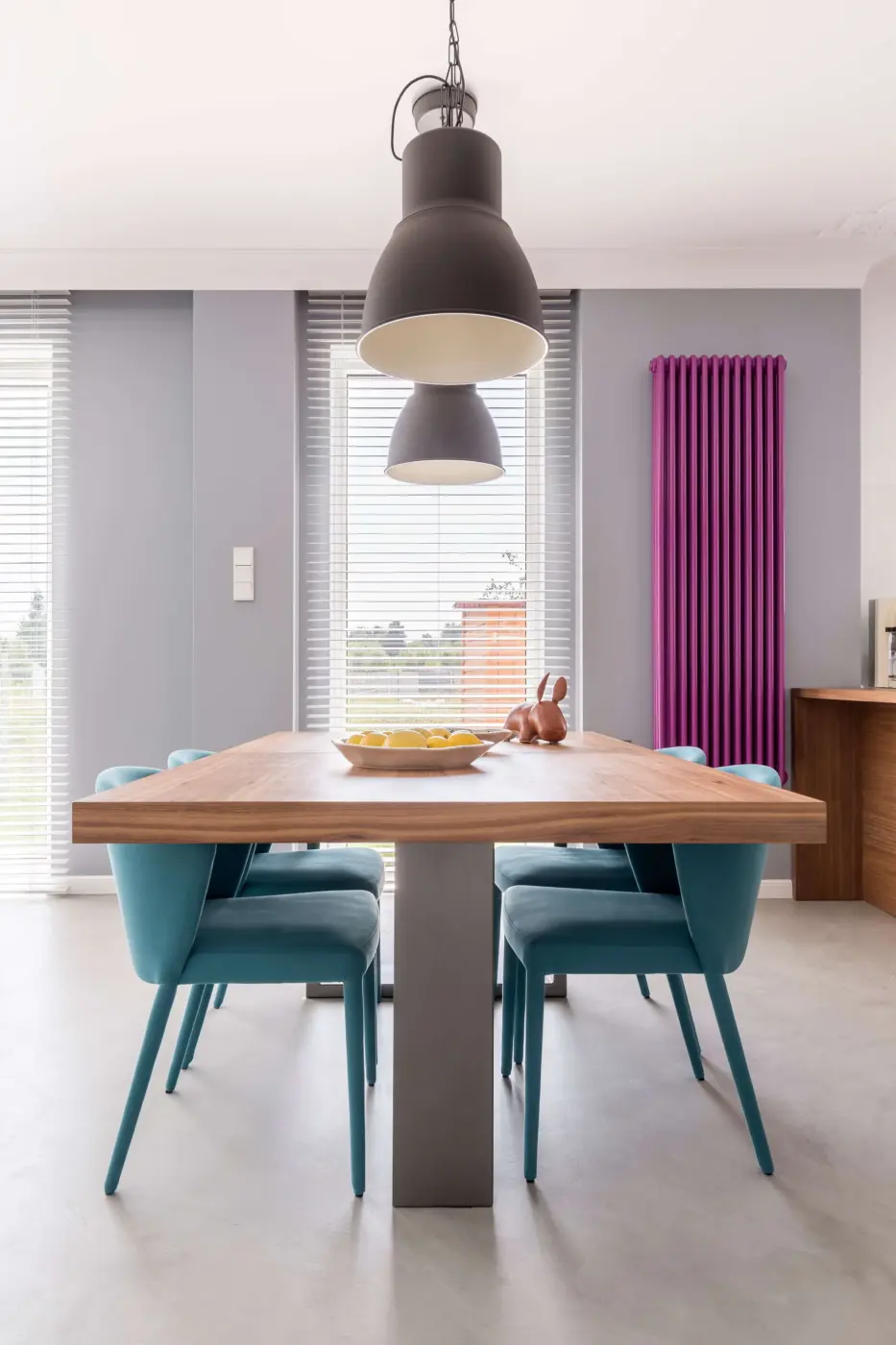 Webdesign Hannover - Ein modernes Esszimmer, fotografiert von immobilien fotograf hannover, zeichnet sich durch einen Holztisch, sechs tealfarbene Stühle, Pendelleuchten, eine Schale mit Zitronen und eine stilvolle Dekoration aus. Graue Wände, hohe Fenster mit Jalousien und ein vertikaler lila Heizkörper vervollständigen den Look. Local SEO Agentur Hannover.